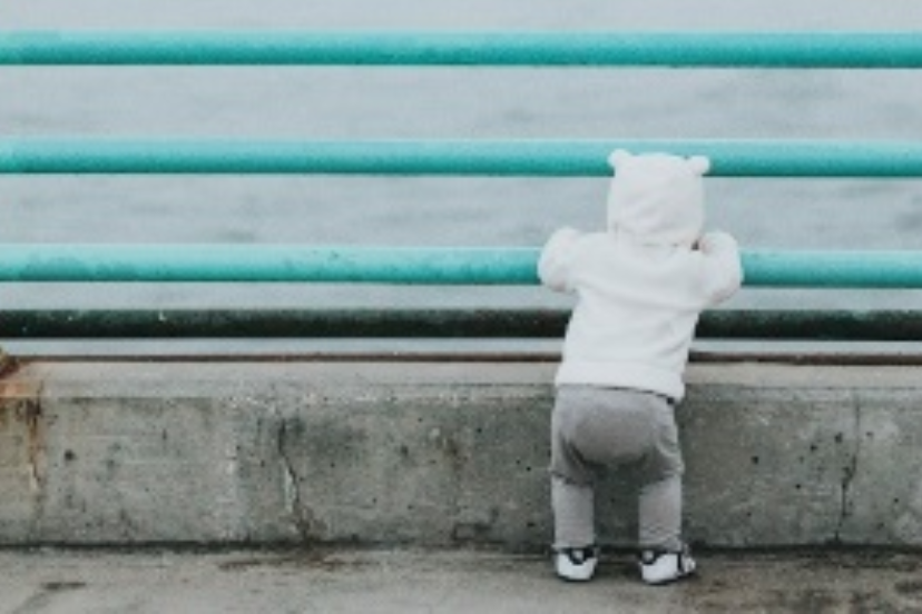 Baby in cute hoodie looking between railings at the ocean.